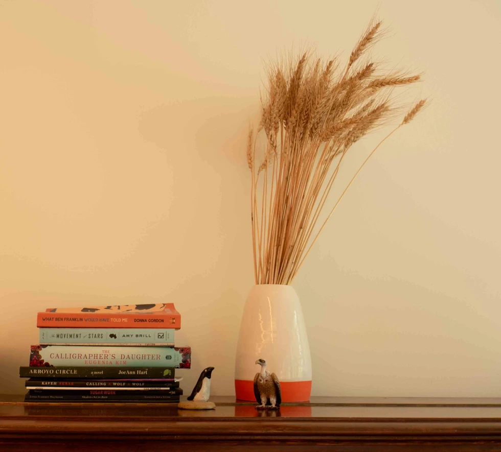 Top of piano with stack of books, vulture figuring, and vase filled with wheat
